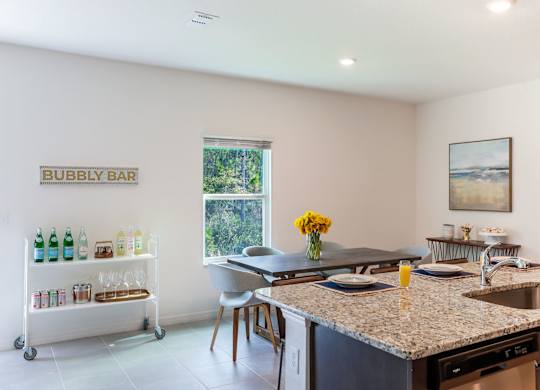 Bright, modern kitchen with granite countertops, dining table set with sunflowers, and a bubbly bar cart.