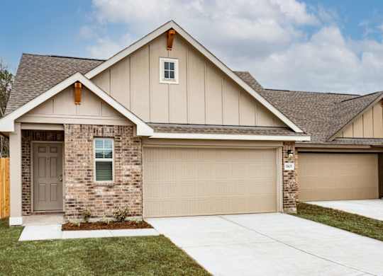 Pet-Friendly Apartments in Humble, TX - Edgewater at the Lagoon - Property Exterior with Brown and Tan Walls, White Trim, Stone Details, Parking Garage, Beige Garage Door, Plant Bed, Grass Lawns, and Concrete Driveway.