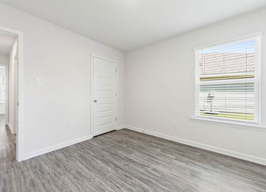 Empty bedroom with gray wood-look flooring, white walls, a window with blinds, and a white door.
