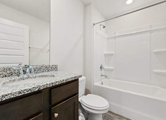 Modern bathroom with granite countertop, dark wood vanity, toilet, and white bathtub-shower combination.