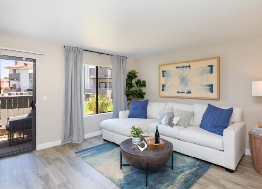 Dog-Friendly Apartments in Moreno Valley, CA - Sorelle Apartments - Living Room with Light-Gray Walls, Wood-Style Floor, Large Windows with Dark-Gray Curtains, Sliding Glass Door to a Patio, White Couch, Brown Kettle-Shaped Coffee Table, Circular Side Table, and Lamp