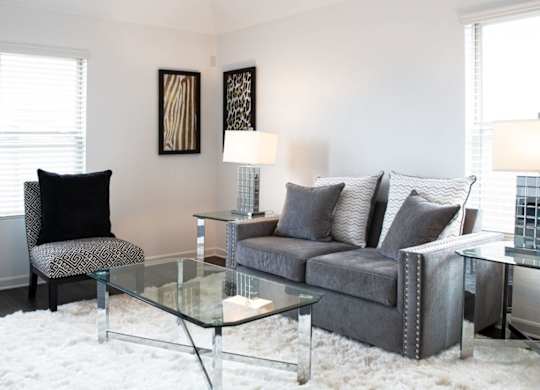 Two-Bedroom Apartments in Humble, TX - Edgewater at the Lagoon - Living Room with Vinyl Plank Flooring, Fluffy White Area Rug, Glass Cofee Table, End Tables with Lamps, Grey Couch, Graphic Cushioned Chair, Two Windows, and Framed Wall Art.