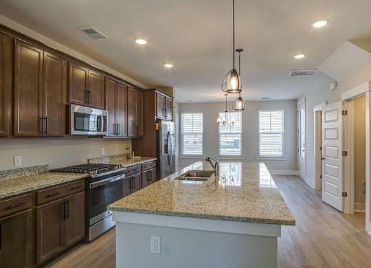 Houses for Rent in North Charleston, SC - Sumner Village - Kitchen with Wood-Style Flooring, Granite Countertops, Stainless Steel Appliances Including a Refrigerator, Oven, and Built-In Microwave, Brown Cabinetry, a Large Kitchen Island, Overhead Hanging Lights, and a View of the Connected Living Area.