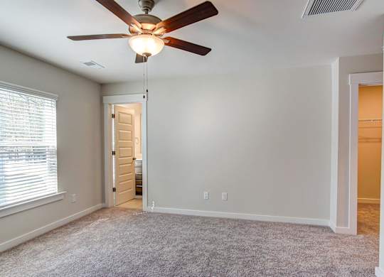 Dog-Friendly Townhomes in Park Circle, SC - Sumner Village - Empty Bedroom with Plush Carpeting, a Ceiling Fan with Lighting, an Attached Bathroom with Tile Flooring, a Spacious Attached Closet, and Large Windows with White Blinds.
