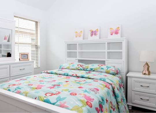 Pet-Friendly Apartments in Humble, TX - Edgewater at the Lagoon - Bedroom with a White-Framed Bed and Bright Bedding, Nightstand with Lamp, White Dresser with Drawers, and Window with White Blinds.