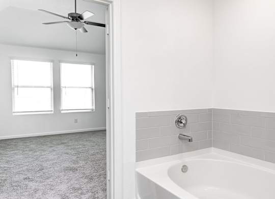 Humble, TX, Apartments for Rent - Edgewater at the Lagoon - Attached Bathroom with Bath Tub, Grey Tile Backsplash, and View of Bedroom with Carpeted Flooring and Ceiling Fan.