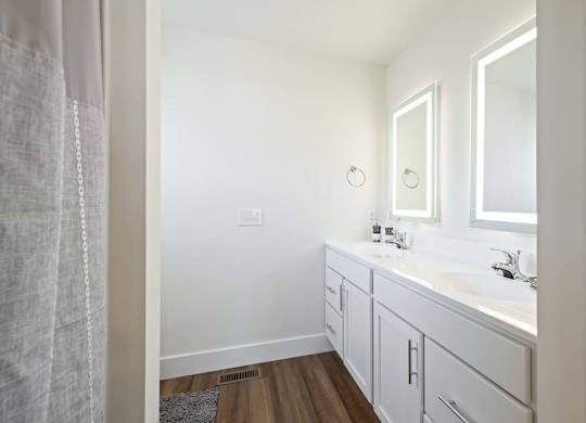 Modern bathroom with dual sinks, white cabinetry, lighted mirrors, and wood-style flooring.