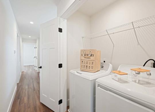 Laundry area with washer, dryer, shelving, and wood-style flooring in a bright, modern hallway.