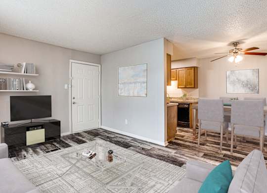 Fort Worth TX Apartments for Rent - Monarch Pass - Living Room with Wood-Style Floor, Beige Walls, Gray Couches, Coffee Table with Clear Glass Top, and a View of the Dining Area and Kitchen