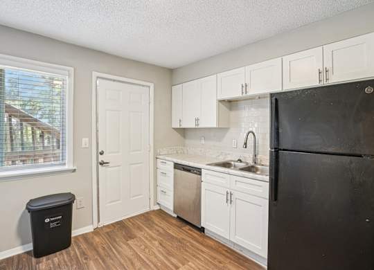 A black refrigerator sits in a kitchen with white cabinets and a window.