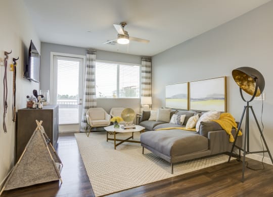 Pet-Friendly Apartments In South Scottsdale, AZ - Lucent Scottsdale - Living Room with Luxurious Wood-Vinyl Plank Flooring, Area Rug, Grey Sectional Couch, White Armchairs, Coffee Table, Side Table with Lamp, Standing Lamp, Wall-Mounted TV, Storage Cabinet, Ceiling Fan, Large Window, and Access to Patio.
