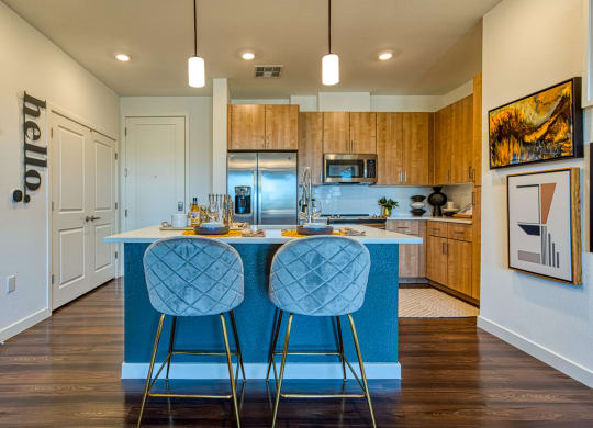 Two-Bedroom Apartments in South Scottsdale, AZ - Lucent Scottsdale – Kitchen with Luxurious Wood-Vinyl Plank Flooring, Area Rug, Chic Island Kitchen with Seating, Pendant Lights, Premium Stainless Steel Appliances, Wood-Tone Cabinetry, Mounted Art, Quartz Countertops, and Ceramic Tile Backsplash.