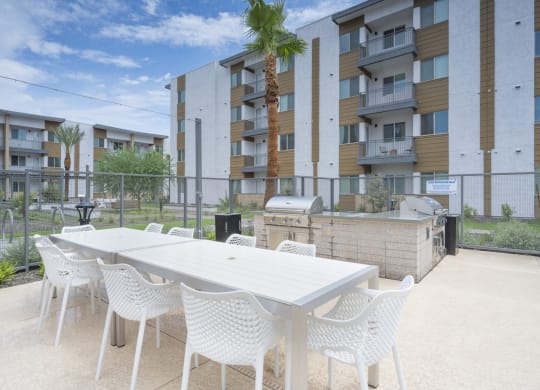 3-Bedroom Apartments In South Scottsdale, AZ - Lucent Scottsdale - BBQ Area With Two  Grilling Stations, Counterspace, Outdoor Dining Tables, Chairs, And View Of Palm Trees And Apartment Buildings.