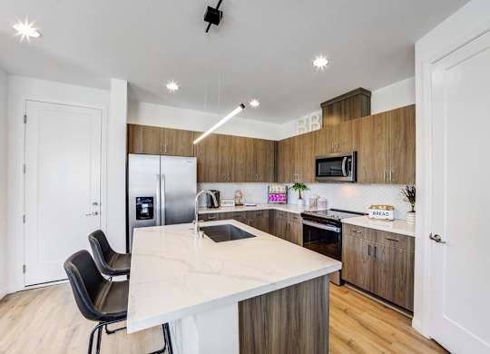 New Luxury Apartments In Skye Canyon, NV - Banyan Brighton - Open-Concept Kitchen With Wood-Inspired Flooring, Marble Countertops, Hardwood Cabinets, Tile Backsplash, Energy Efficient Appliances, A Kitchen Island, And A Pantry.