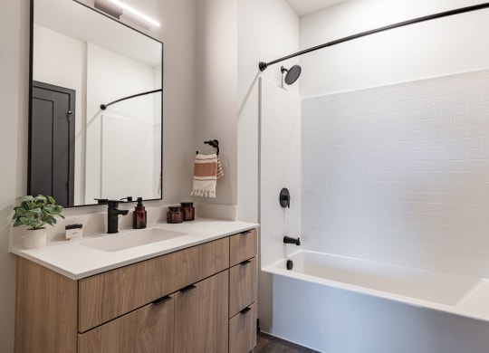 Bloomington, MN Apartments Near Mall of America - Carbon31 - Modern Bathroom with White Walls, Shower and Bathtub Combo, Sink, White Countertop, Black Plumbing Fixtures, and Large Mirror
