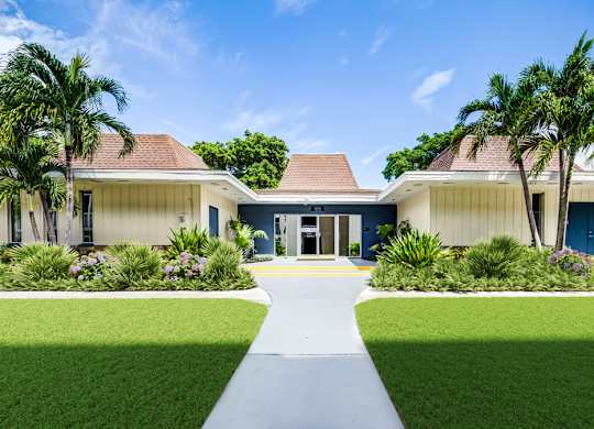 Entrance to the leasing office surrounded by palm trees and landscaped lawns at Coronado Springs Apartments in Palm Springs, FL. Pet-friendly community with one- and two-bedroom apartments.