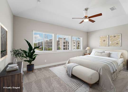 A bedroom with a large bed, a ceiling fan, and a view of the city through the windows.