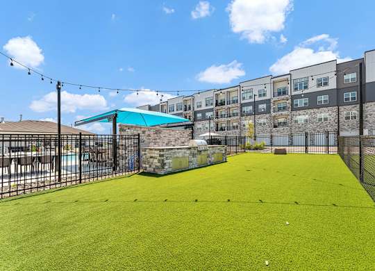 A green artificial turf soccer field with a fence and a building in the background.