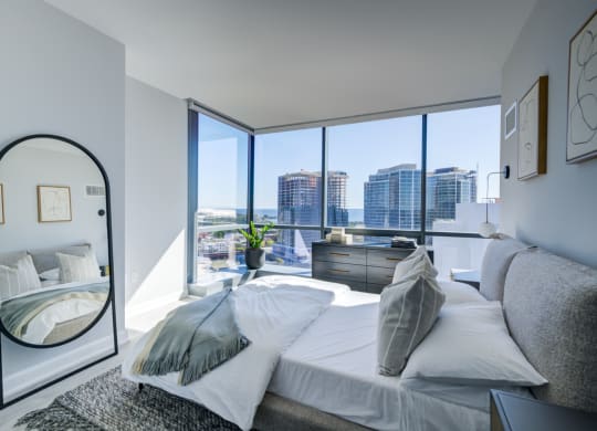 A modern bedroom featuring a plush bed with grey bedding, a large floor mirror, and a panoramic view of the city skyline through floor-to-ceiling windows