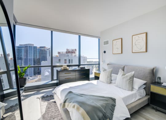 Bright and airy city-view bedroom with a comfortable bed adorned with white and grey pillows, flanked by minimalist nightstands and abstract wall art.