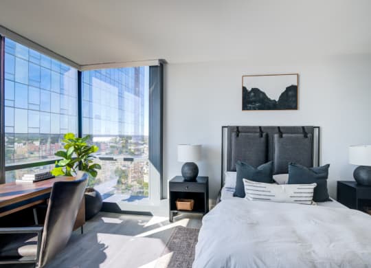 Spacious bedroom with a king-sized bed, crisp white linens, and contrasting black accent pillows, complete with a work desk overlooking an expansive urban landscape.