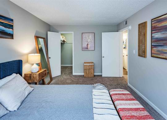 A bedroom with a bed, a nightstand with a lamp, a mirror, a doorway leading to another room, and a painting on the wall.