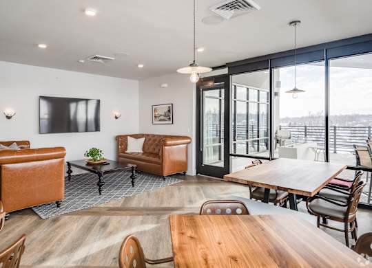 One Bedroom Luxury Apartments in Greeley, CO - The Shelby - Resident Clubroom with Wood-Style Flooring, Two Leather-Style Couches, Wall-Mounted TV, Multiple Wood-Style Tables with Chairs, Overhead Hanging Lights, and Floor-to-Ceiling Windows with View of the Outdoor Patio Lounge.