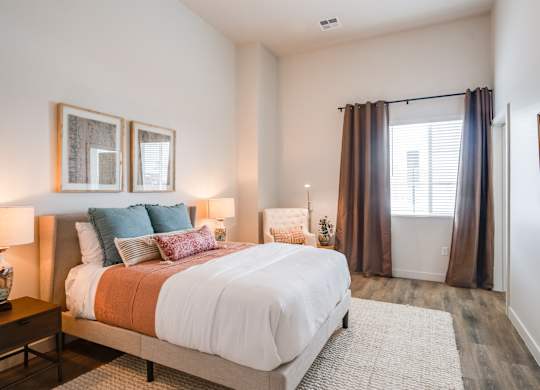 Luxury Apartments in Greeley, CO, with In-Unit Washer and Dryer - The Shelby - Bedroom with Wood-Style Flooring, a High Ceiling, a Bed, Two Bedside Tables with Lamps, and Window with Blinds and Curtains.