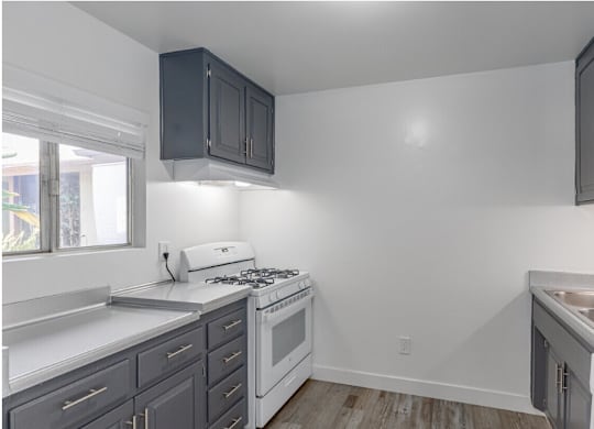 El Cajon, CA Apartments - Ivanhoe Village Apartments - Compact Kitchen With Gray Cabinetry, White Gas Range, Double Sink, Light Countertops, And Wood Style Flooring.