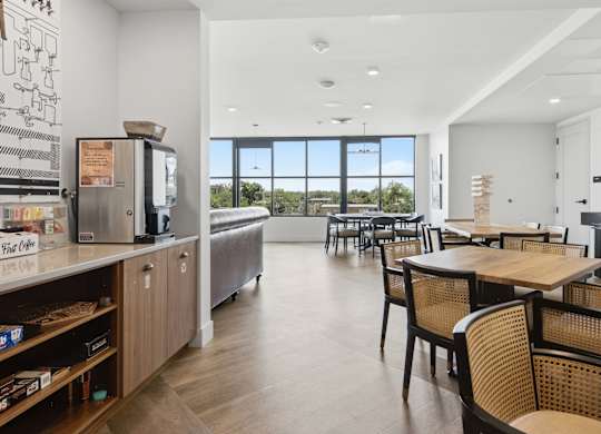 A kitchen with a refrigerator, a dining table with chairs, and a view of the outdoors through the windows.