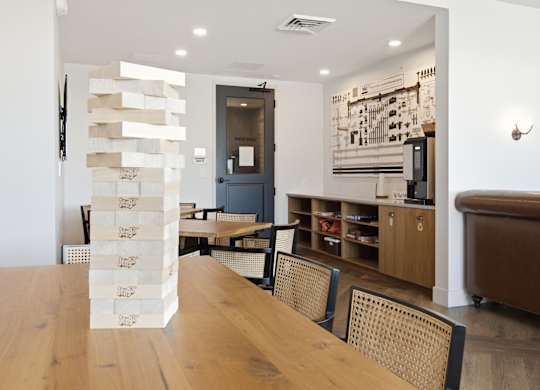 A room with a wooden table and chairs and a white wall with a picture on it.