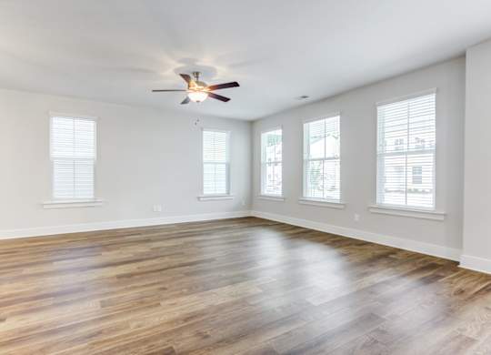 Single Family Homes in North Charleston, SC - Sumner Village - Empty Living Room Area with Hardwood-Style Flooring, a Ceiling Fan with Lighting, and Multiple Large Windows with White Blinds.