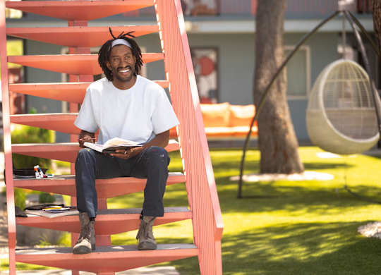 A man with dreadlocks is sitting on a red staircase reading a book.