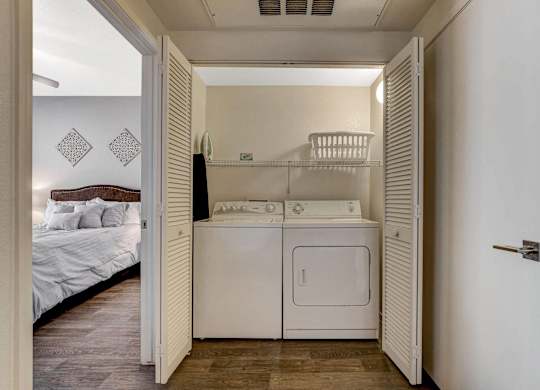 A white laundry machine is in a small room with a bed in the background.