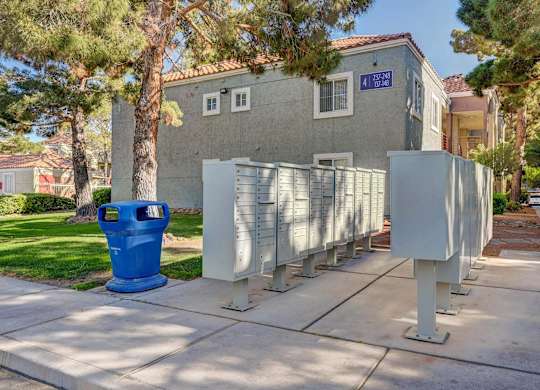 A blue trash can is in front of a grey building with a tree in front of it.