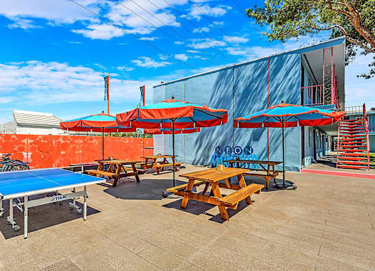 A blue ping pong table is surrounded by red umbrellas and wooden picnic tables.