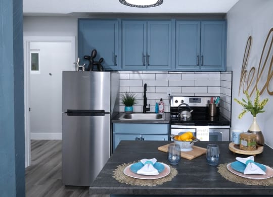 Fusion Las Vegas efficiency apartments in Las Vegas Nevada kitchen with stainless steel appliances, navy blue cabinets, subway tile backsplash, and two place settings on island.