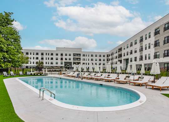 A large swimming pool surrounded by sun loungers and umbrellas in front of a modern building.