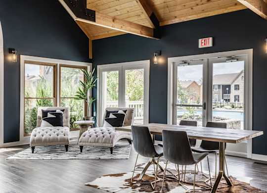 A community clubhouse featuring two cozy comfy chairs, a table surrounded by chairs, hardwood-style flooring, and large windows.