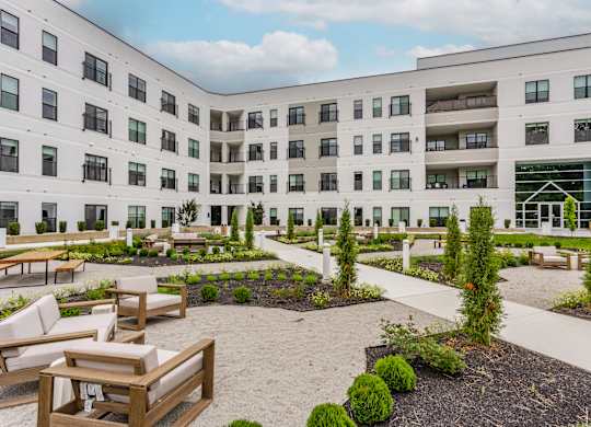 A courtyard with a white building in the background and a few trees and chairs in the foreground.