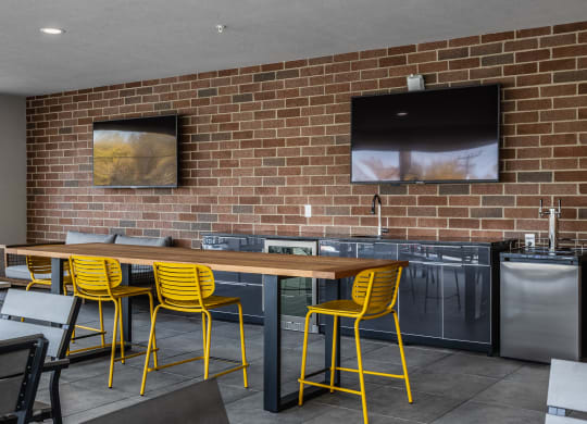 A sky lounge with a large table surrounded by four yellow chairs, next to two TV's on a brick wall.