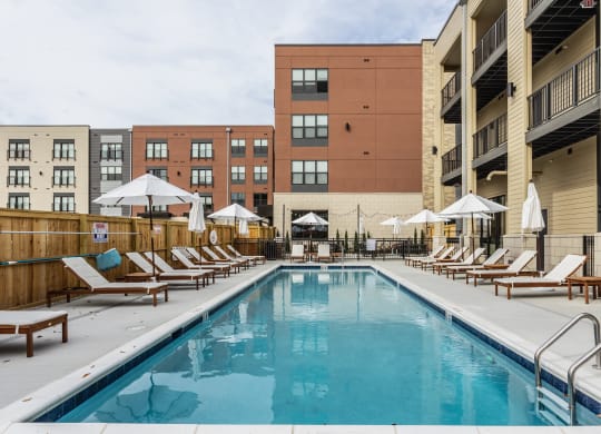 A swimming pool surrounded by a fence, white chairs and white umbrellas, next to apartment buildings.
