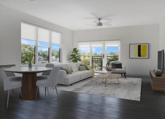 A spacious living room with a white couch and chair, a rug on hardwood-style floors, and large windows, next to a dining room table.