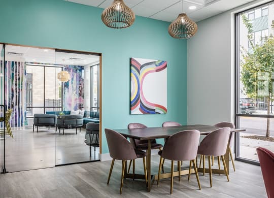 A co-working lounge at Madamore with a blue wall, a table surrounded by six chairs, and a large window, on hardwood-style floors.