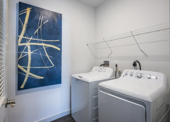 A closet with a shelf, a white washer and dryer, and a painting on the wall.