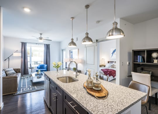 Kitchen featuring an island with a sink and granite countertops, next to a living room with a large window.