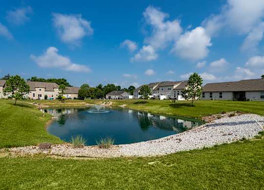 A pond in the middle of a grassy area with houses in the background.