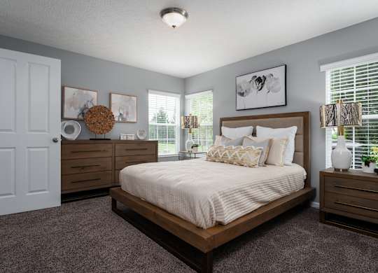 Large carpeted bedroom at Carson Farms with a bed, nightstands, dresser, lamps, and three bright windows