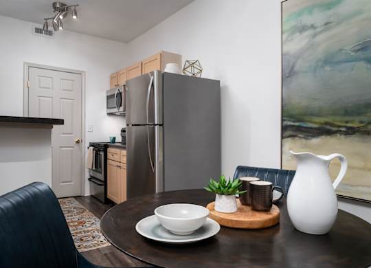 A modern kitchen with a refrigerator, a dining table with a bowl, a plant, and a jug.