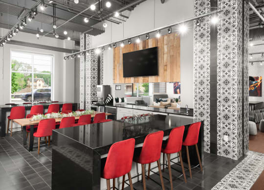 A seating area with tables surrounded by red chairs, and  light fixtures, next to a kitchen and a TV.